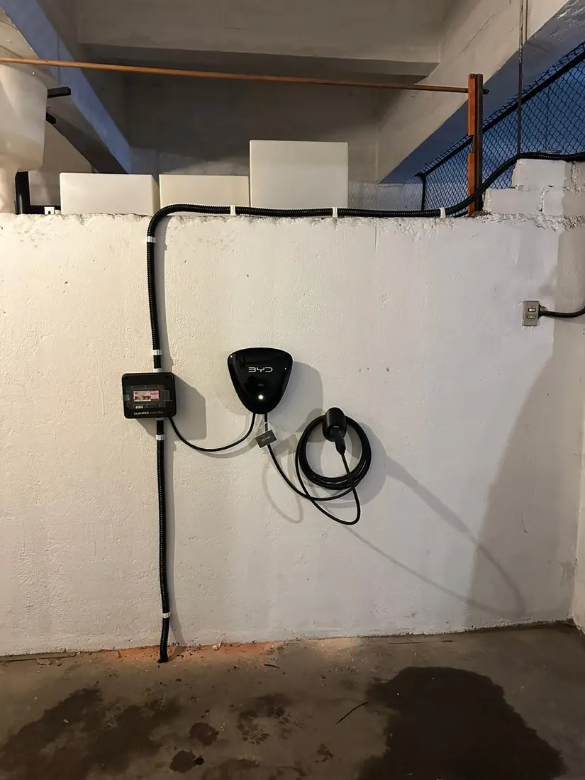 Carregador BYD preto fixado em parede de garagem, ligado a um quadro de proteção Clamper por eletroduto corrugado
